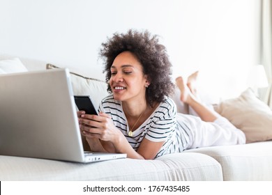 Happy Young Black Woman Using Mobile Phone While Sitting A Couch At Home With Laptop Computer. Smiling Woman Sitting On Sofa Relaxing While Browsing Online Shopping Website.