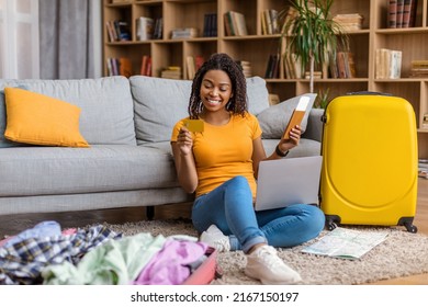 Happy Young Black Woman Using Credit Card And Laptop, Booking Abroad Trip Or Making Hotel Reservation On Web, Holding Passport With Tickets, Sitting Near Suitcases At Home