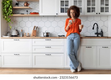 Happy Young Black Woman Using Smartphone And Drinking Morning Coffee, Smiling African American Female Standing In Kitchen Interior, Messaging With Friends And Enjoying Hot Drink At Home, Full Length - Powered by Shutterstock