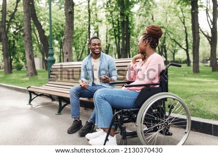 Happy young black woman in wheelchair talking to her boyfriend at park, free space. Happy African American guy sitting on bench, communicating to his female friend with disability outdoors