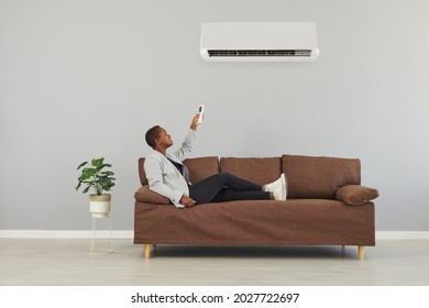 Happy Young Black Woman Who's Lounging On Comfortable Brown Sofa In Minimalist Home Interior Looks Above At Air Conditioner And Presses Power Switch On Remote Control. Modern AC System Concept