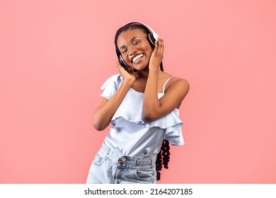 Happy Young Black Woman In Wireless Headphones Listening To Popular Music On Pink Studio Background. Cheerful African American Female Enjoying Favorite Song Or Cool Playlist