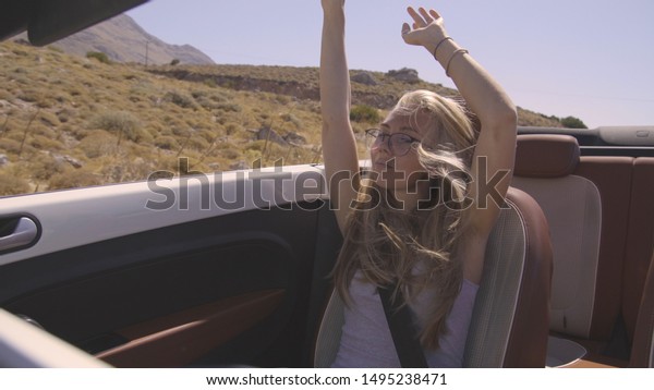 Happy young blond woman enjoying a white\
convertible car ride. Pretty blonde girl riding in cabriolet with\
raised hands on a warm summer day. Windy hair,travel,freedom,escape\
and destination concept.