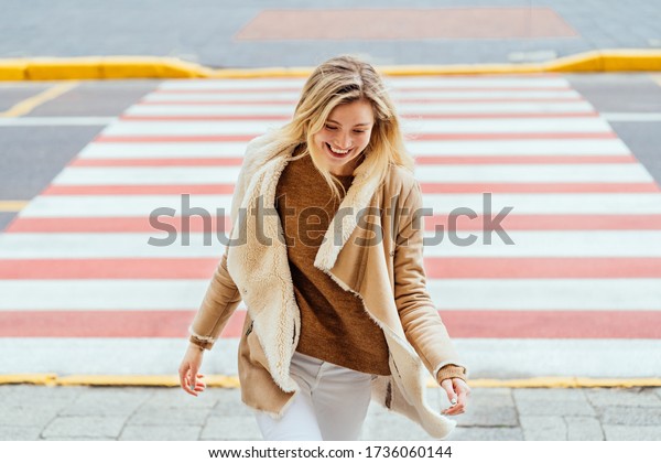 Happy young blond woman go fast, walking on\
pedestrian crossing on street while looking down and smiling.\
Confident woman wearing casual\
clothes.