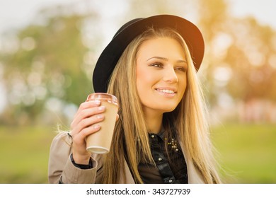 Happy Young Blonde Caucasian Woman In Park In Autumn Wearing Trench Coat And Fedora Hat Holding A Cup Of Takeaway Coffee. Horizontal, Retouched, Vibrant Colors.