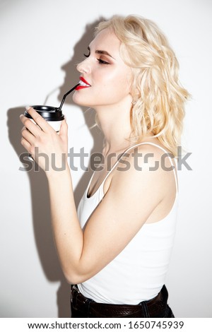 happy young blonde girl drinks from a paper cup through a straw on a white background, studio fashion photo