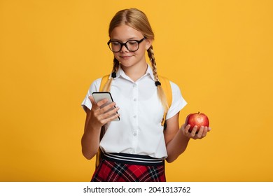 Happy Young Blonde Girl Pupil With Pigtails And Backpack In Glasses Typing On Phone, Holds Red Apple, Isolated On Yellow Background. Health Care, Proper Nutrition, Lifestyle, Education At School