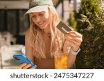 Happy young blonde woman with cap customer shopper holding credit card using cell phone mobile app buying fashion clothes paying online make purchase in ecommerce digital store on smartphone in cafe.