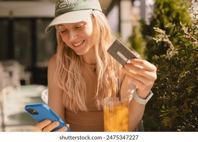 Happy young blonde woman with cap customer shopper holding credit card using cell phone mobile app buying fashion clothes paying online make purchase in ecommerce digital store on smartphone in cafe. - Powered by Shutterstock