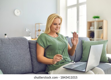 Happy Young Blonde Woman Sitting On Couch At Home And Waving Hand At Laptop Computer Starting Video Call. Concepts Of Teaching And Tutoring Online, E-learning, Remote Work And Social Distancing