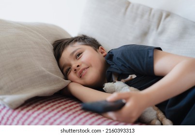 Happy Young Boy Holding Remote Control And Looking Up With Smiling Face, Cute Kid Lying On Sofa Enjoy Watching TV Program, Child Relaxing At Home On Weekend