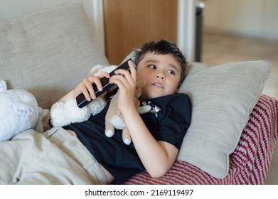 Happy Young Boy Holding Remote Control And Looking Up With Smiling Face, Cute Kid Lying On Sofa Enjoy Watching TV Program, Child Relaxing At Home On Weekend