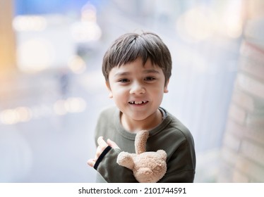Happy young boy looking up with smiling face holding teddy bear standing next to window glass with bright light shining,Positve Kid playing with soft toy brown bear, Child relaxing at home in morning 