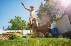 Happy Young Boy Play Jump Over Water Sprinkler Lifting Up Hands In The Garden Lawn During Summer Hot Day