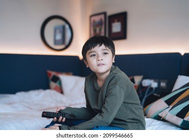 Happy Young Boy Sitting In Bed Holding Remote Control, Cute Kid Looking Out With Smiling Face, Child Relaxing At Home Watching TV In Bed Room.