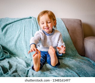 Happy Young Boy Wearing Blue Jeans And White Sweater Sits On Grey Couch With Ocean Blue Throw Blanket Counting His Toes.