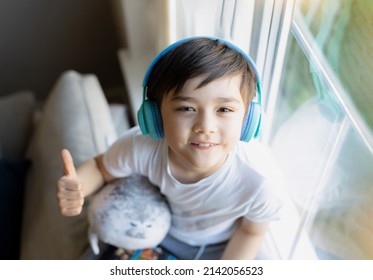 Happy Young Boy Wearing Headphones Listening To Music. Cute Kid Looking Up To Camera With Smiling Face, Positive Child Sitting Next To Window Relaxing On Weekend In Spring Or Summertime