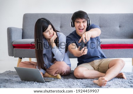 Happy young boyfriend wear headphone holding joystick playing video game with make fun and tease girlfriend to displeased and angry in living room at home. Asian young couple leisure vacation concept.