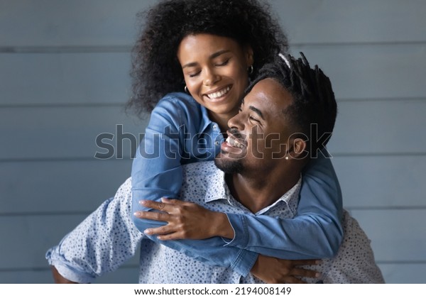 Happy young Brazilian ethnicity man giving\
piggyback ride to laughing beautiful African American wife, having\
fun together near grey wall, good trusted family relations,\
entertainment activity