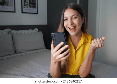 Happy young Brazilian woman holding smartphone and celebrates with a clenched fist sitting on bed at home - Powered by Shutterstock