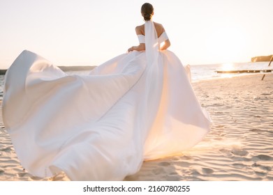 Happy young bride woman in white dress running, have fun on clean sandy beach waves of a sea or ocean on sunset, summer shooting near a water. Wedding rest, relax honeymoon concept.