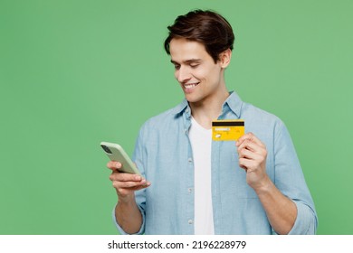 Happy Young Brunet Man 20s Years Old Wears Blue Shirt Using Mobile Cell Phone Hold Credit Bank Card Doing Online Shopping Order Delivery Booking Tour Isolated On Plain Green Background Studio Portrait