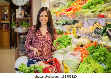 Happy Young Brunette Carrying A Shopping Basket And Picking Some Vegetables At The Grocery Store