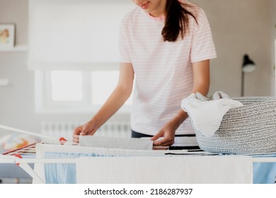 Happy young brunette caucasian woman smiling, hanging clean wet clothes laundry on drying rack at home. Female housewife hands closeup spreading laundry from basket. Girl puts towels after drying them