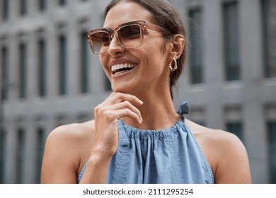 Happy young brunette European woman holds chin looks away smiles broadly being in good mood wears trendy sunglasses and fashionable clothes poses against blurred background spends free time in city - Shutterstock ID 2111295254
