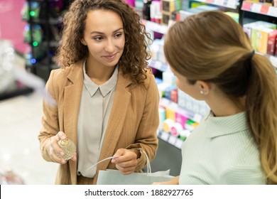 Happy young brunette female with bottle of scent going to apply perfume on paper blotter while testing new fragrance with friend in beauty shop