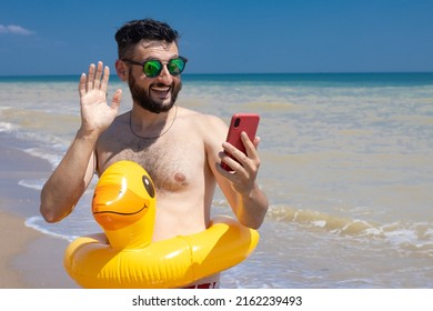 Happy Young Brunette Man In Sunglasses Making Video Call With Smartphone, Gesturing Hi To Friend Or Relative, Wearing Yellow Duck Inflatable Ring Standing On Sea Beach On Summer Sunny Day.Copy Space