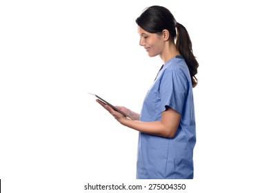 Happy Young Brunette Nurse Or Doctor In Blue Uniform Smiling While Using A Thin Wireless PC Tablet Connected To Internet, Side Portrait Isolated On White