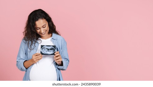 Happy young brunette pregnant woman with big tummy showing ultrasound image of her baby, holding fetus photo over her chest, pink studio background, panorama with copy space - Powered by Shutterstock