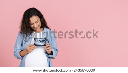 Happy young brunette pregnant woman with big tummy showing ultrasound image of her baby, holding fetus photo over her chest, pink studio background, panorama with copy space