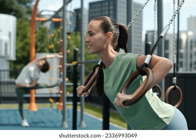 Happy young brunette sportswoman hanging on still rings on sports ground while working out against another woman doing exercise - Powered by Shutterstock