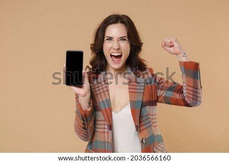 Happy young brunette woman 20s in casual checkered jacket hold mobile cell phone with blank empty screen mock up copy space doing winner gesture isolated on beige colour background, studio portrait