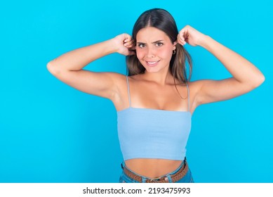 Happy Young Brunette Woman Wearing Blue Tank Top Over Blue Background Ignores Loud Music And Plugs Ears With Fingers Asks To Turn Off Sound