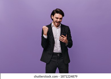 Happy young brunette-haired man in white shirt and black striped suit emotionally looking at phone, rejoicing and posing against violet pastel background