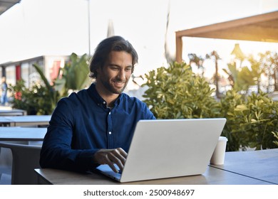 Happy young business man entrepreneur using laptop sitting outdoors with coffee. Smiling guy student or professional worker looking at computer in cafe elearning, hybrid working, searching job online. - Powered by Shutterstock