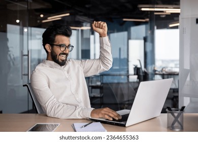 Happy young business man working in the office at the laptop, sitting at the table and rejoicing in success, raised his smiling hand up as a sign of victory. - Shutterstock ID 2364655363