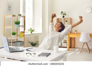 Happy young business woman sitting at office desk with eyes closed, stretching arms and back trying to relax stiff, tense muscles, enjoying break during working day