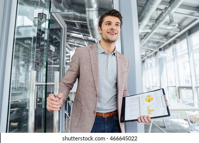 Happy Young Businessman With Business Plan On Clipboard Entering The Door In Office