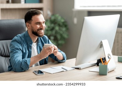 Happy Young Businessman Drinking Coffee At Workplace And Using Computer, Handsome Male Entrepreneur Sitting At Work Desk, Looking At Monitor Screen And Smiling, Enjoying Casual Day In Office - Powered by Shutterstock