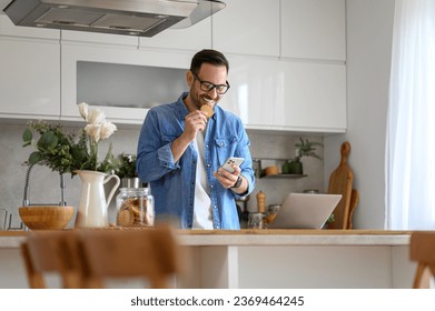 Happy young businessman eating cookie while checking messages over mobile phone at kitchen counter - Powered by Shutterstock