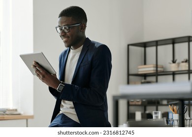 Happy young businessman in elegant suit looking through online financial data on tablet screen while standing in front of camera in office