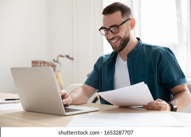 Happy Young Businessman In Eyeglasses Working On Computer At Modern Office. Smiling Company Employee In Eyeglasses Holding Paper Documents, Chatting Online With Clients On Laptop At Workplace.