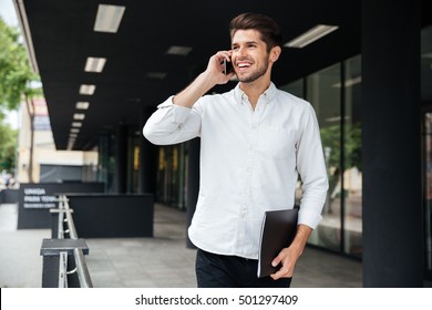 Happy young businessman with folder walking and talking on cell phone near business center - Powered by Shutterstock