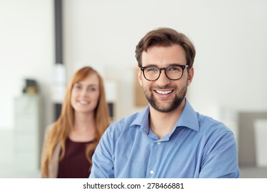 Happy Young Businessman Inside The Office, Wearing Blue Polo Shirt With Eyeglasses, Smiling At The Camera In Front His Female Co-worker.