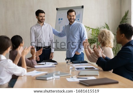 Happy young businessman introduce new male employee newcomer at office meeting, man boss congratulate worker with promotion or achievement, colleagues applaud, acknowledgment concept