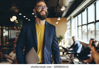 Happy young businessman smiling in a co-working space. Contemplative businessman looking away thoughtfully while holding a laptop. Cheerful young entrepreneur working in a modern workspace. - Shutterstock ID 2090850115